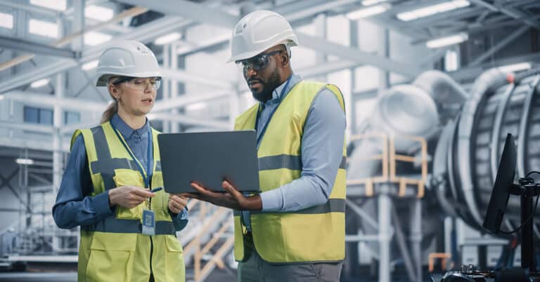 Two diverse technicians/engineers in safety uniforms, hard hats, and glasses, working on a laptop in a factory.